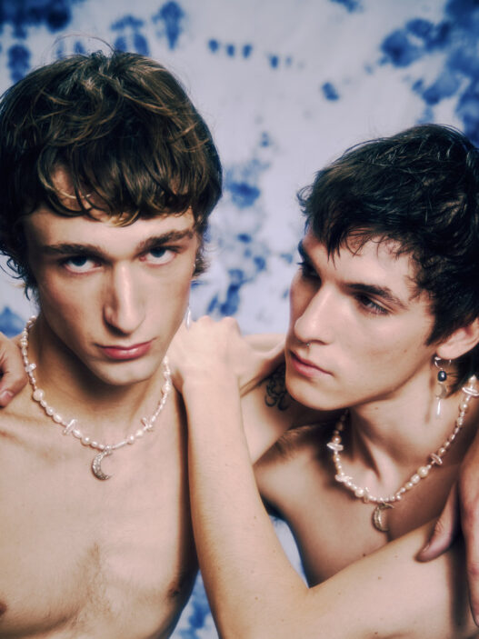Two shirtless young men wearing pearl necklaces lean close, one with an arm around the other, against a blue-dyed backdrop.