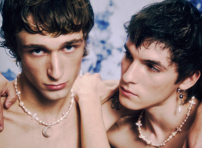 Two shirtless young men wearing pearl necklaces lean close, one with an arm around the other, against a blue-dyed backdrop.