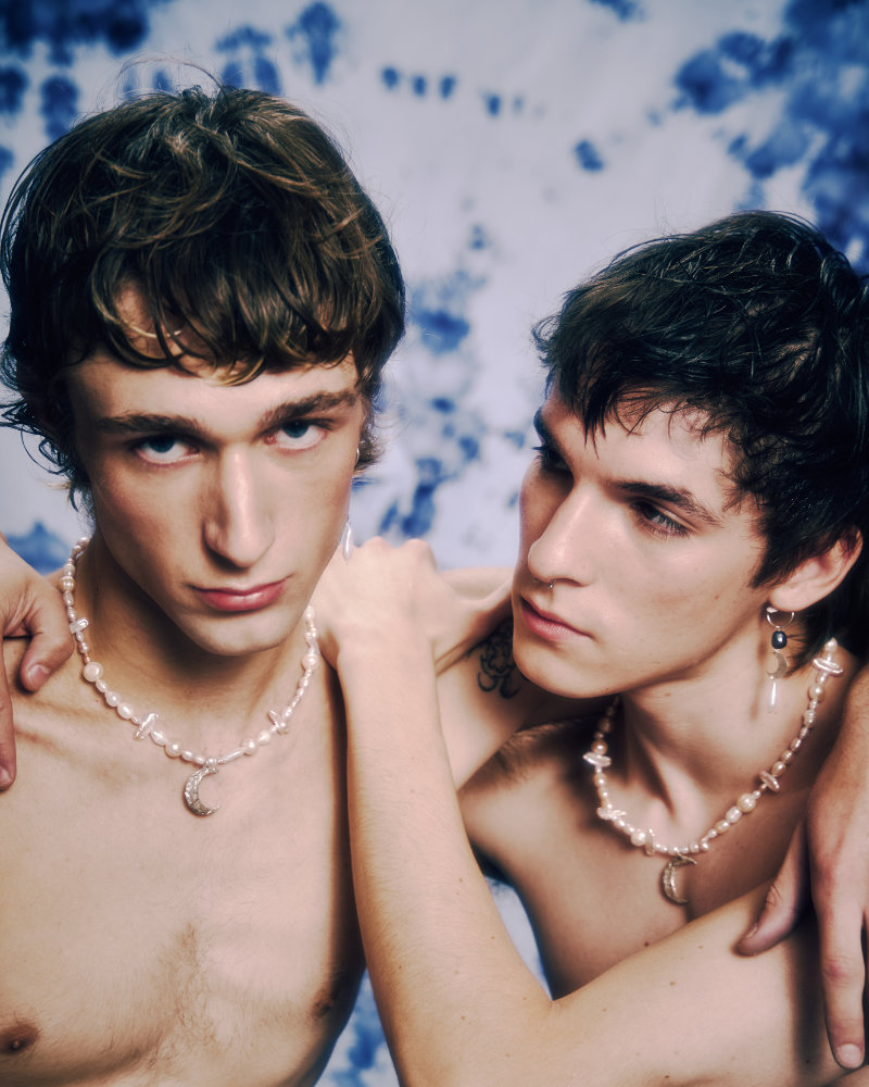 Two shirtless young men wearing pearl necklaces lean close, one with an arm around the other, against a blue-dyed backdrop.