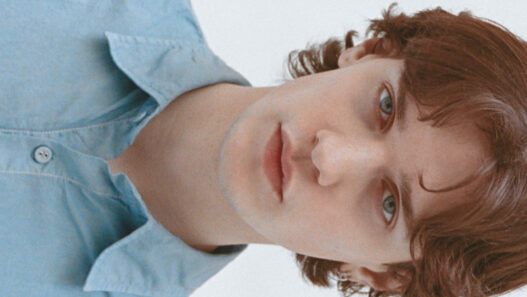 Portrait of a young person with wavy brown hair wearing a light blue button-up shirt, looking at the camera.
