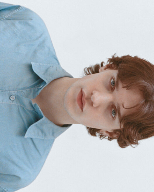 Portrait of a young person with wavy brown hair wearing a light blue button-up shirt, looking at the camera.