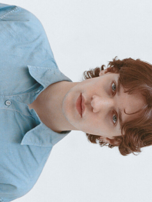 Portrait of a young person with wavy brown hair wearing a light blue button-up shirt, looking at the camera.