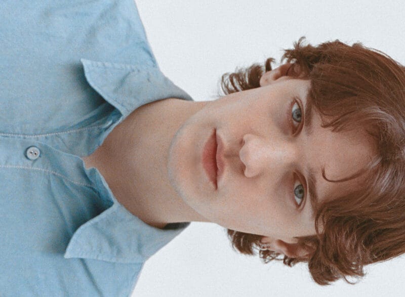Portrait of a young person with wavy brown hair wearing a light blue button-up shirt, looking at the camera.