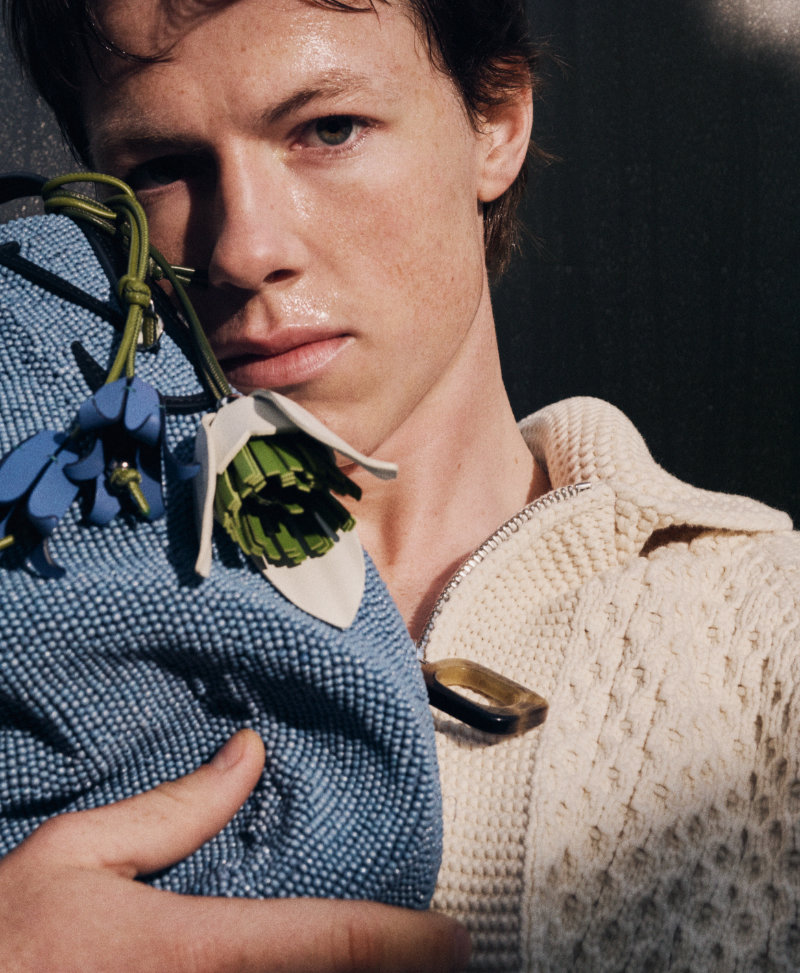Close-up portrait of a person in a cream knit sweater holding a blue woven bag with blue floral embellishments.