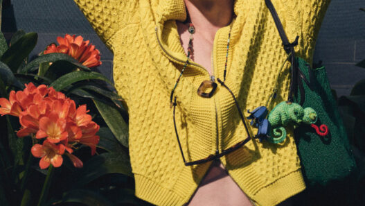 Young person in a bright yellow knit sweater, hands behind the head, sunglasses hanging from the neck, with orange tropical flowers and green plants in the background.