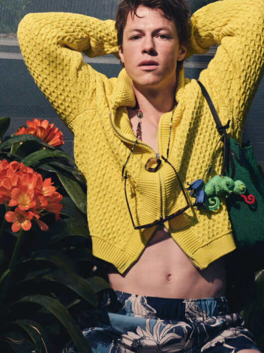 Young person in a bright yellow knit sweater, hands behind the head, sunglasses hanging from the neck, with orange tropical flowers and green plants in the background.