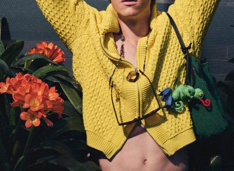 Young person in a bright yellow knit sweater, hands behind the head, sunglasses hanging from the neck, with orange tropical flowers and green plants in the background.