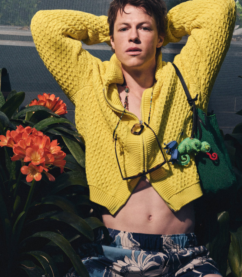 Young person in a bright yellow knit sweater, hands behind the head, sunglasses hanging from the neck, with orange tropical flowers and green plants in the background.