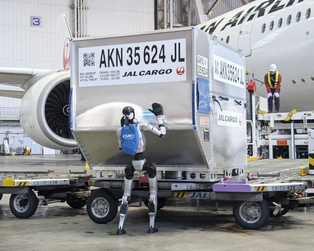 A humanoid robot stands beside a large silver JAL Cargo container on a baggage cart in a hangar, waving at the container with a blue backpack, with a jet engine and a worker in a high‑visibility vest in the background.