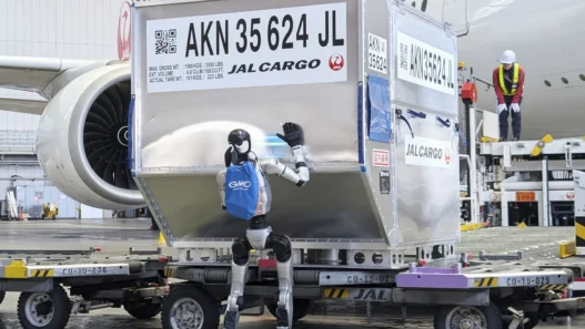 A humanoid robot stands beside a large silver JAL Cargo container on a baggage cart in a hangar, waving at the container with a blue backpack, with a jet engine and a worker in a high‑visibility vest in the background.