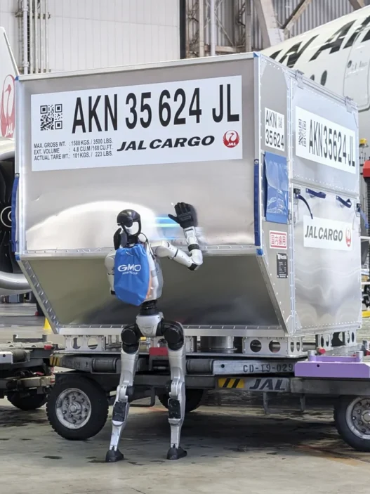 A humanoid robot stands beside a large silver JAL Cargo container on a baggage cart in a hangar, waving at the container with a blue backpack, with a jet engine and a worker in a high‑visibility vest in the background.