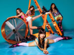 Four women in colorful outfits and roller skates pose around an orange ladder and a large industrial fan in a bright studio with a blue background.