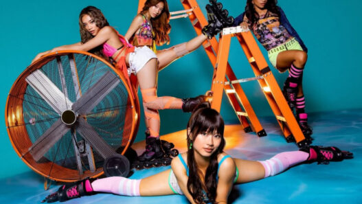 Four women in colorful outfits and roller skates pose around an orange ladder and a large industrial fan in a bright studio with a blue background.