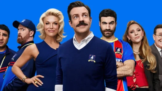 Ensemble cast posing in a row against a solid blue background, center man in navy sweater smiles at camera.