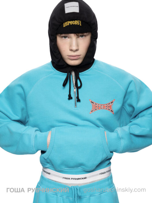 Model wearing a turquoise hoodie and pants with a black hooded balaclava; branded chest logo visible.