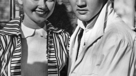 Black-and-white photo of a smiling woman with short curled hair standing beside a young man with dark, slicked-back hair outside a brick building; they hold hands in front of them.