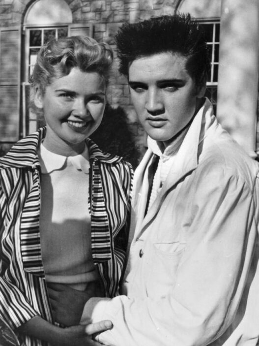 Black-and-white photo of a smiling woman with short curled hair standing beside a young man with dark, slicked-back hair outside a brick building; they hold hands in front of them.