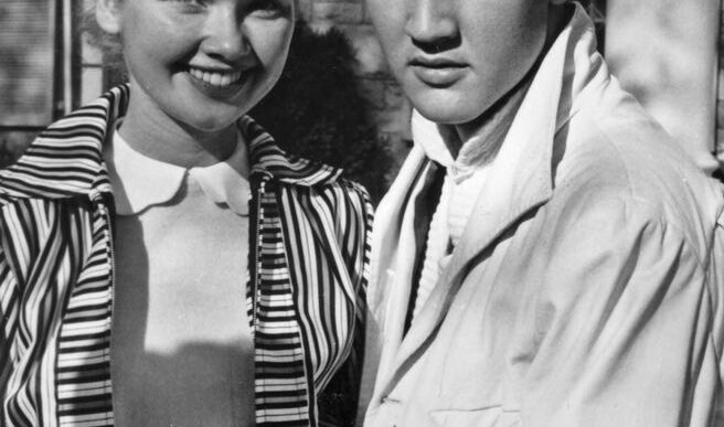 Black-and-white photo of a smiling woman with short curled hair standing beside a young man with dark, slicked-back hair outside a brick building; they hold hands in front of them.
