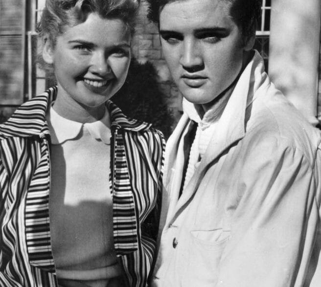 Black-and-white photo of a smiling woman with short curled hair standing beside a young man with dark, slicked-back hair outside a brick building; they hold hands in front of them.