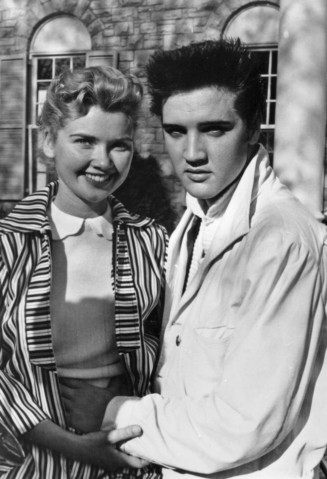 Black-and-white photo of a smiling woman with short curled hair standing beside a young man with dark, slicked-back hair outside a brick building; they hold hands in front of them.