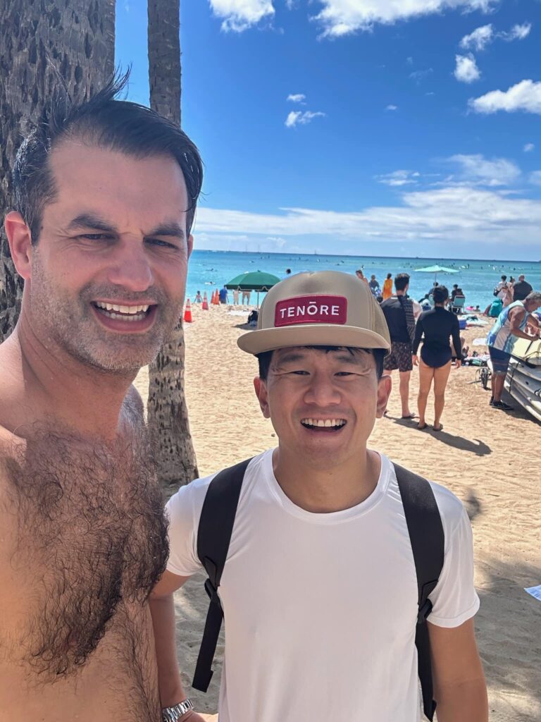 Two men take a beach selfie, smiling, with a palm tree and blue ocean in the background.