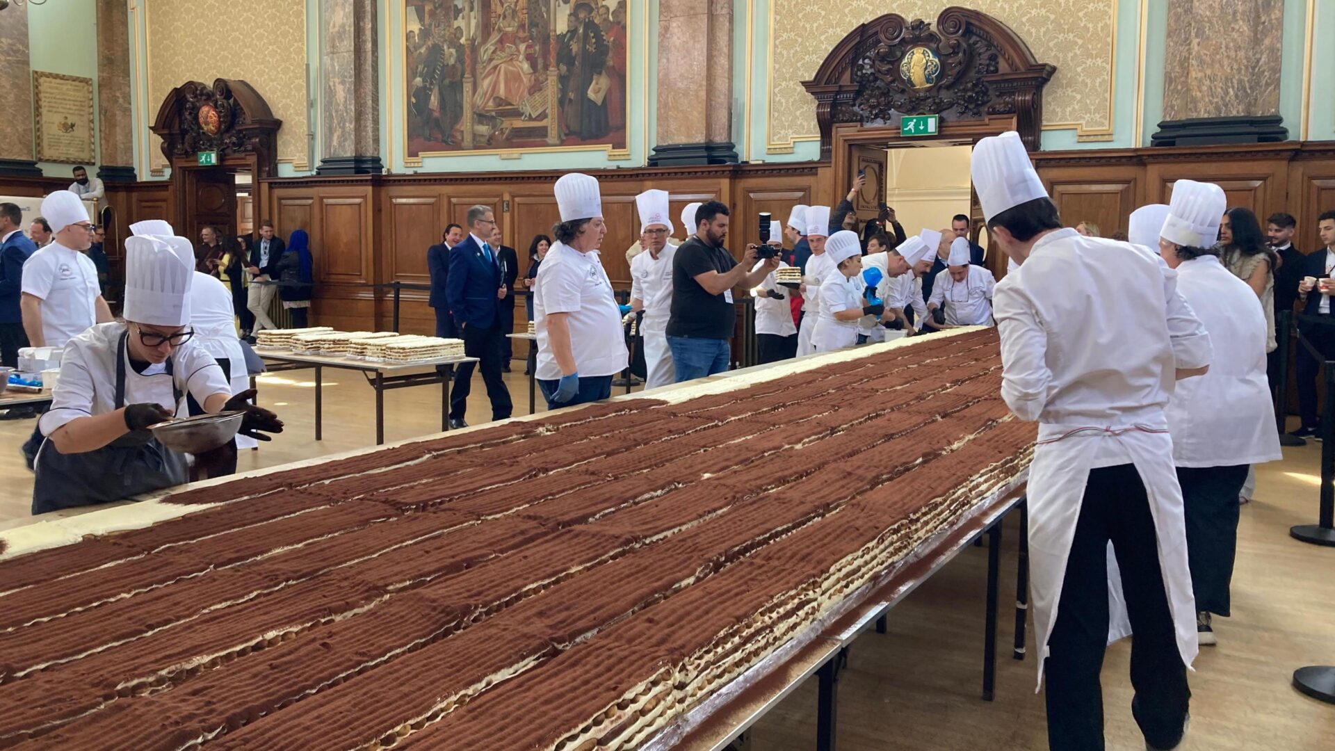 Long row of pastry chefs in white uniforms and tall hats assemble thin chocolate cakes in a grand hall as onlookers watch from the background.