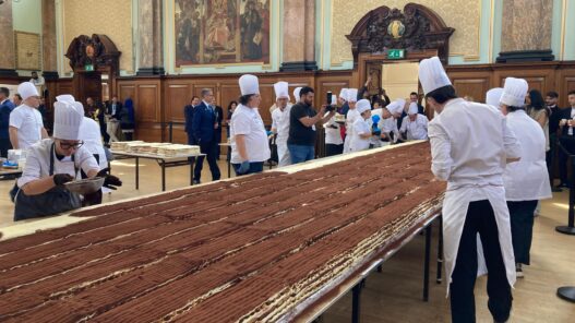 Long row of pastry chefs in white uniforms and tall hats assemble thin chocolate cakes in a grand hall as onlookers watch from the background.