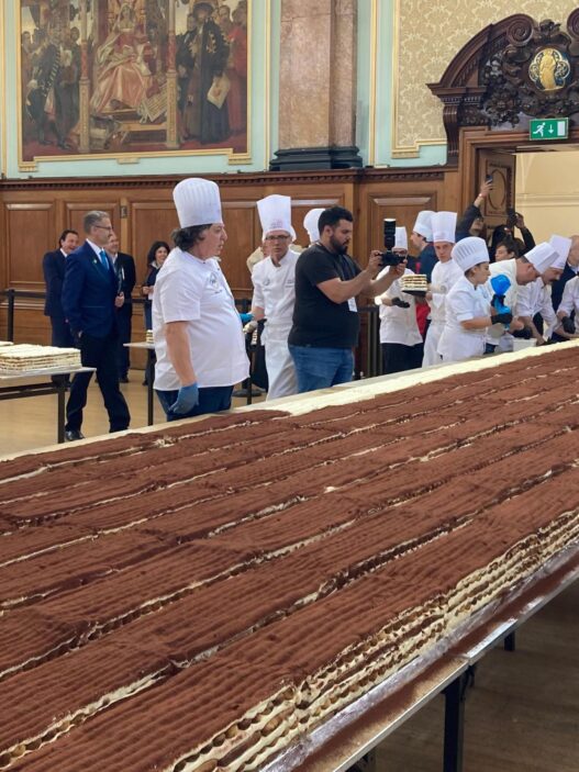 Long row of pastry chefs in white uniforms and tall hats assemble thin chocolate cakes in a grand hall as onlookers watch from the background.