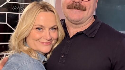 Smiling blonde woman and a man with glasses and a mustache hug in front of a pink wall and glass door.