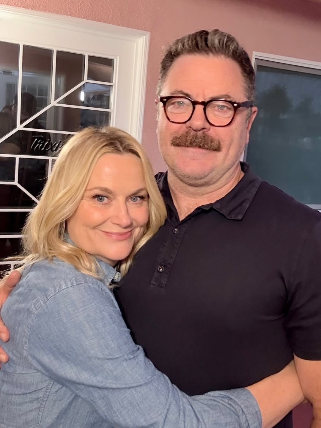 Smiling blonde woman and a man with glasses and a mustache hug in front of a pink wall and glass door.