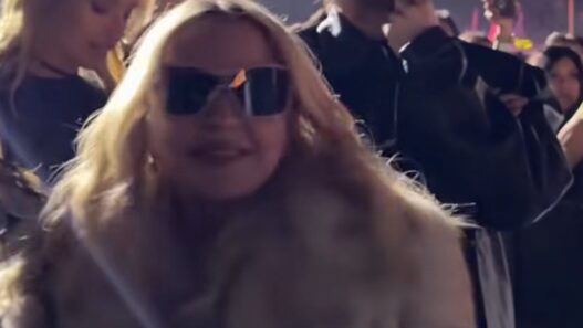 Crowd at a nighttime event; woman in front wearing sunglasses and a fur coat, man behind with sunglasses and hand on chin.