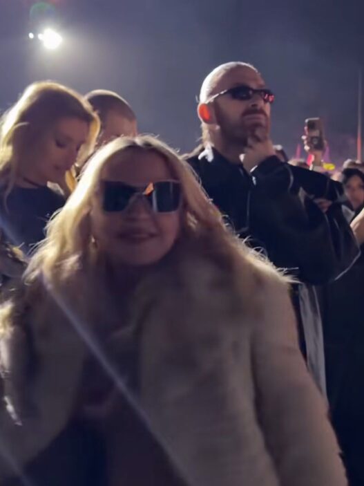 Crowd at a nighttime event; woman in front wearing sunglasses and a fur coat, man behind with sunglasses and hand on chin.