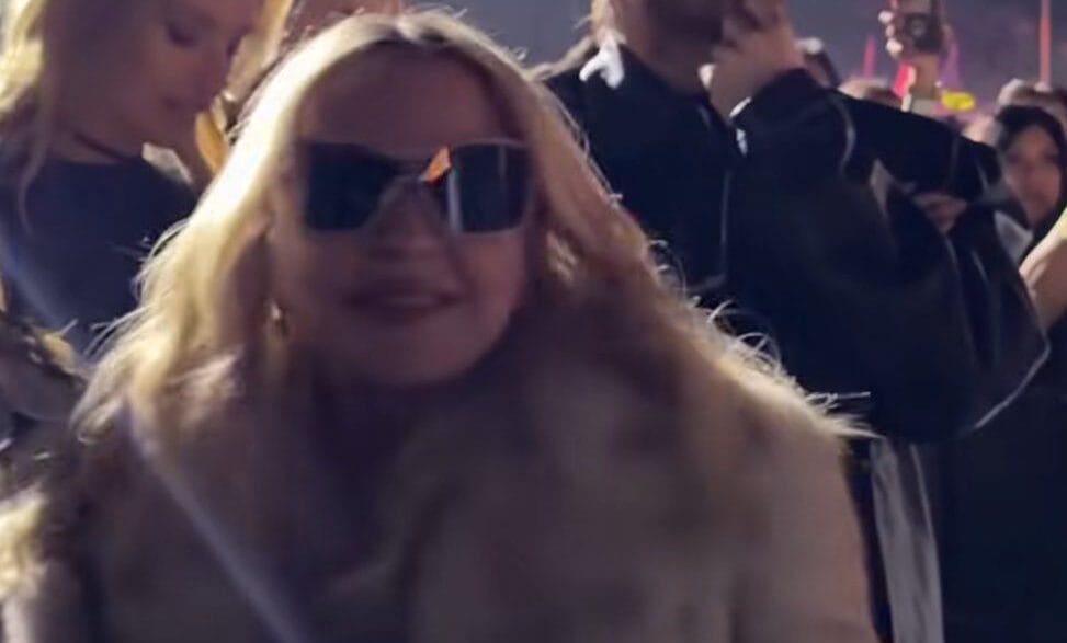 Crowd at a nighttime event; woman in front wearing sunglasses and a fur coat, man behind with sunglasses and hand on chin.
