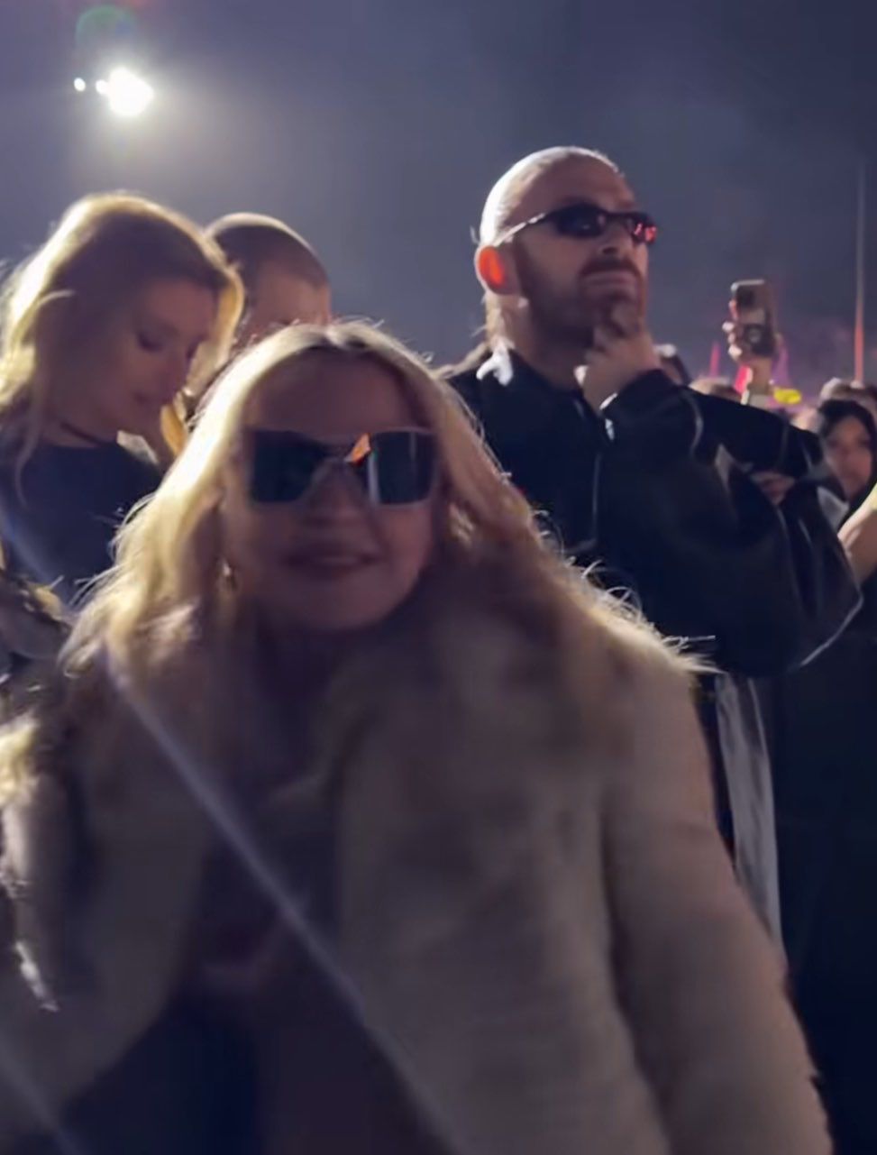 Crowd at a nighttime event; woman in front wearing sunglasses and a fur coat, man behind with sunglasses and hand on chin.