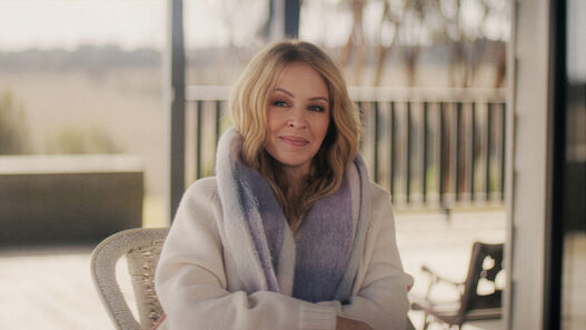 Portrait of a blonde woman in a cream sweater and lavender scarf, sitting on a patio and smiling at the camera.