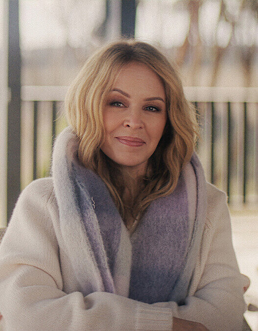 Portrait of a blonde woman in a cream sweater and lavender scarf, sitting on a patio and smiling at the camera.