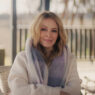 Portrait of a blonde woman in a cream sweater and lavender scarf, sitting on a patio and smiling at the camera.
