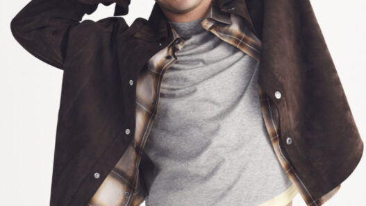 Young man smiling with arms behind his head, wearing a brown suede jacket over a gray T‑shirt and plaid shirt with a white background.