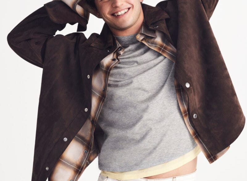 Young man smiling with arms behind his head, wearing a brown suede jacket over a gray T‑shirt and plaid shirt with a white background.