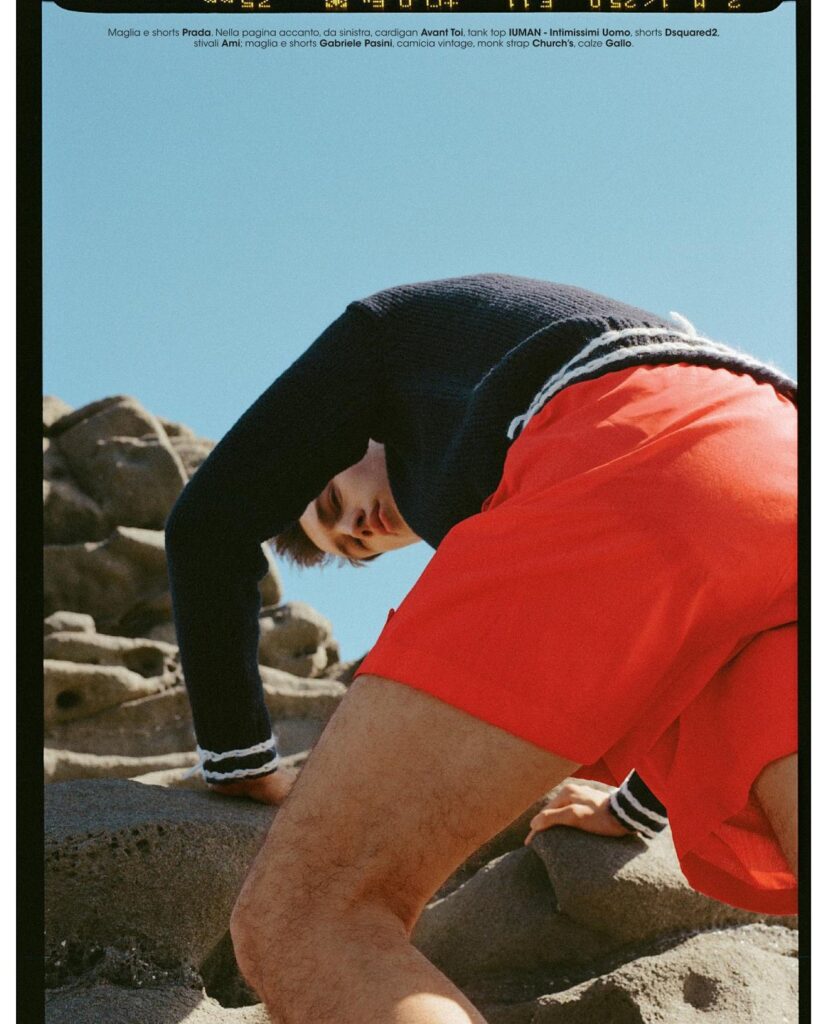Person in a black long-sleeve top and bright red shorts crawls over rocky terrain under a clear blue sky.