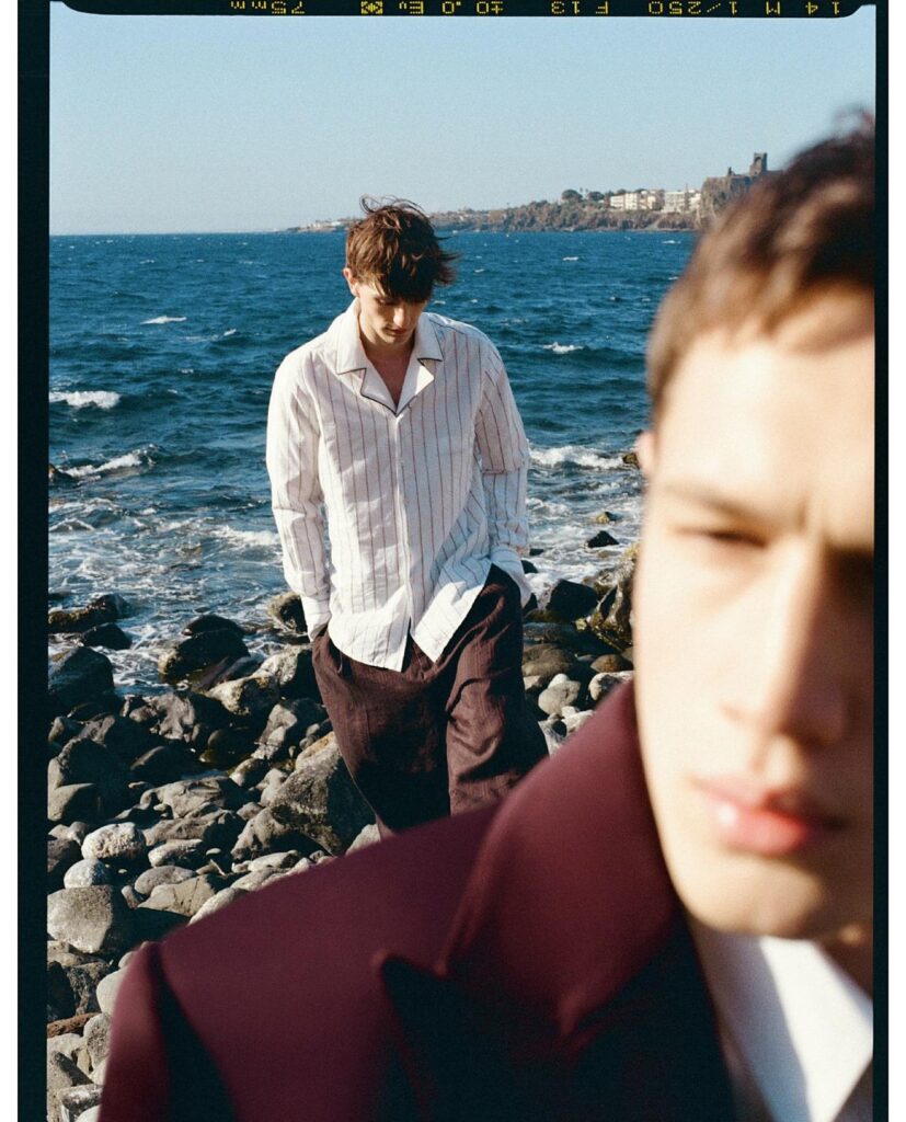 Two young men on a rocky seaside shore; the man in a striped shirt stands by the waves while another figure is partially in the foreground (out of focus).