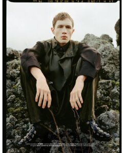 Young man in dark, oversized coat sits on mossy rocks outdoors, looking at the camera with a neutral expression; fashion editorial shot.