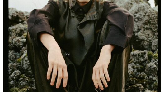 Young man in dark, oversized coat sits on mossy rocks outdoors, looking at the camera with a neutral expression; fashion editorial shot.