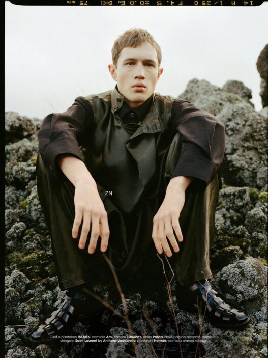 Young man in dark, oversized coat sits on mossy rocks outdoors, looking at the camera with a neutral expression; fashion editorial shot.