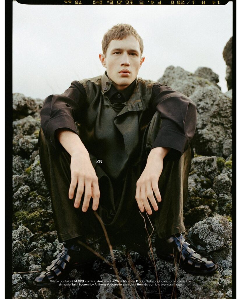Young man in dark, oversized coat sits on mossy rocks outdoors, looking at the camera with a neutral expression; fashion editorial shot.