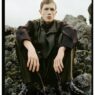 Young man in dark, oversized coat sits on mossy rocks outdoors, looking at the camera with a neutral expression; fashion editorial shot.