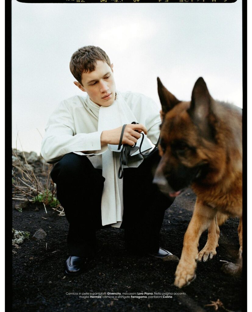 A man in a white shirt crouches to tie a leash on a German Shepherd outdoors on dark soil.