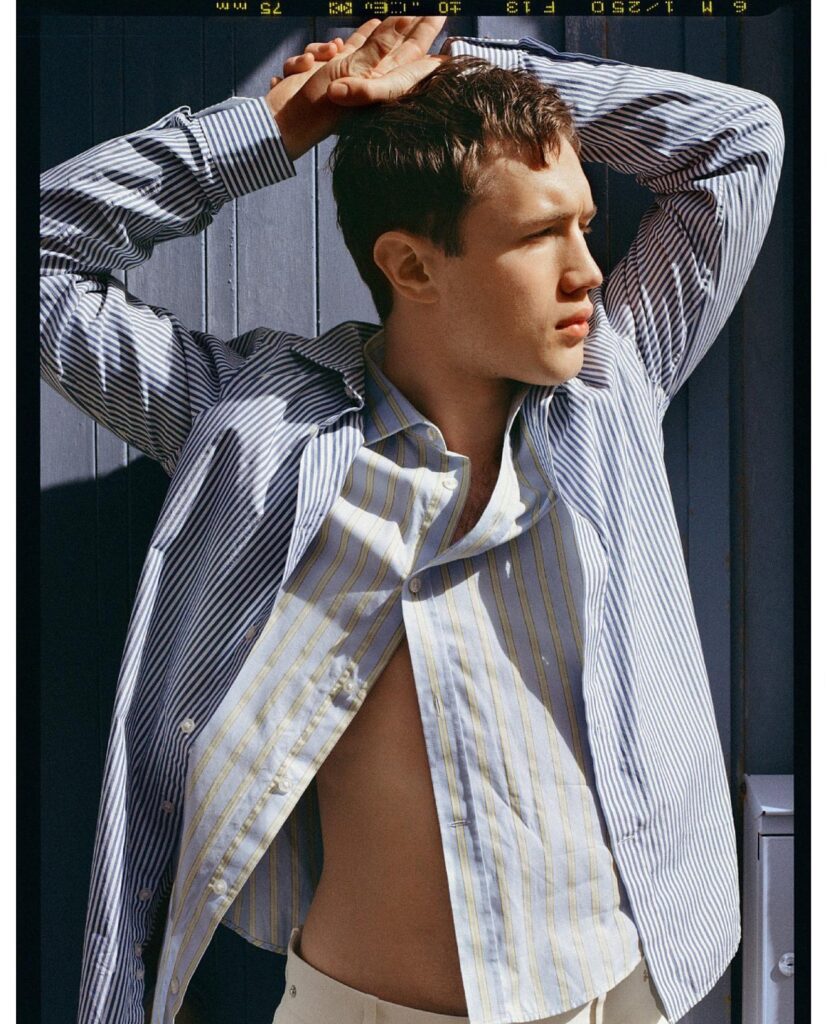 Man wearing a light striped shirt with arms raised, looking to the right against a blue wooden wall.