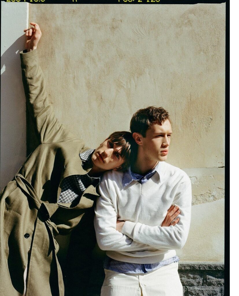 Two young men leaning against a sunlit textured wall; one in a tan trench coat with arm raised, the other in a white sweater.