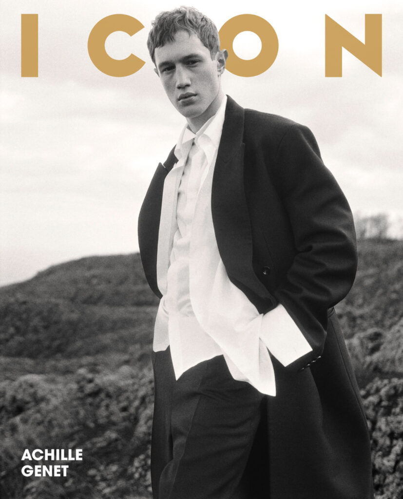 Black-and-white portrait of a young man in a suit with an open white shirt, standing on a rocky landscape; 'ACHILLE GENET' text in the lower left.